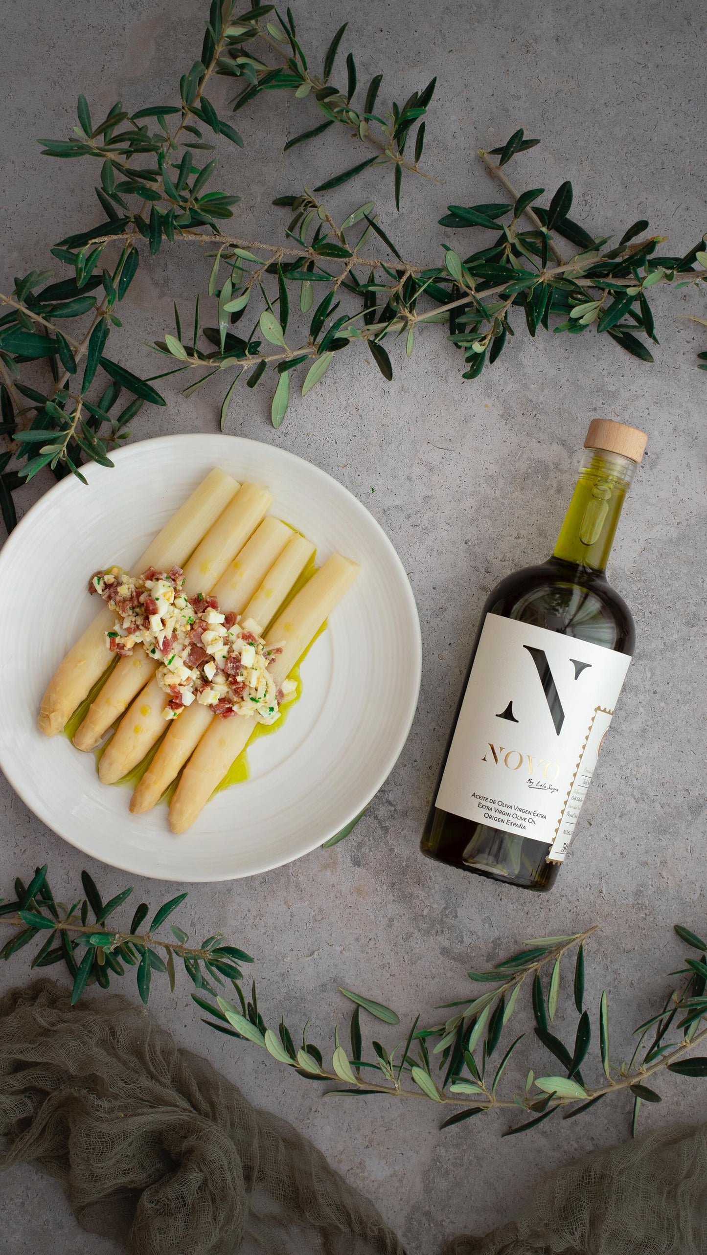 Nobleza del Sur NOVO olive oil bottle with white asparagus dish and decorative olive branches on grey surface