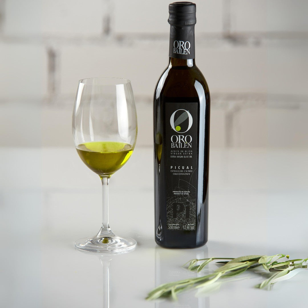 Oro Bailen Picual premium olive oil bottle with wine glass containing golden-green oil and fresh herbs on white surface