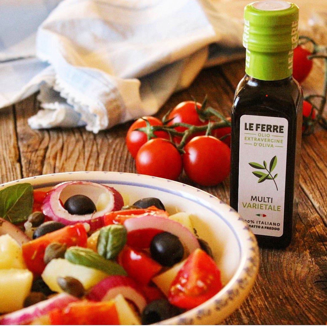LeFerre Multivarietale olive oil bottle next to fresh Mediterranean salad with tomatoes, olives and onions on wooden table