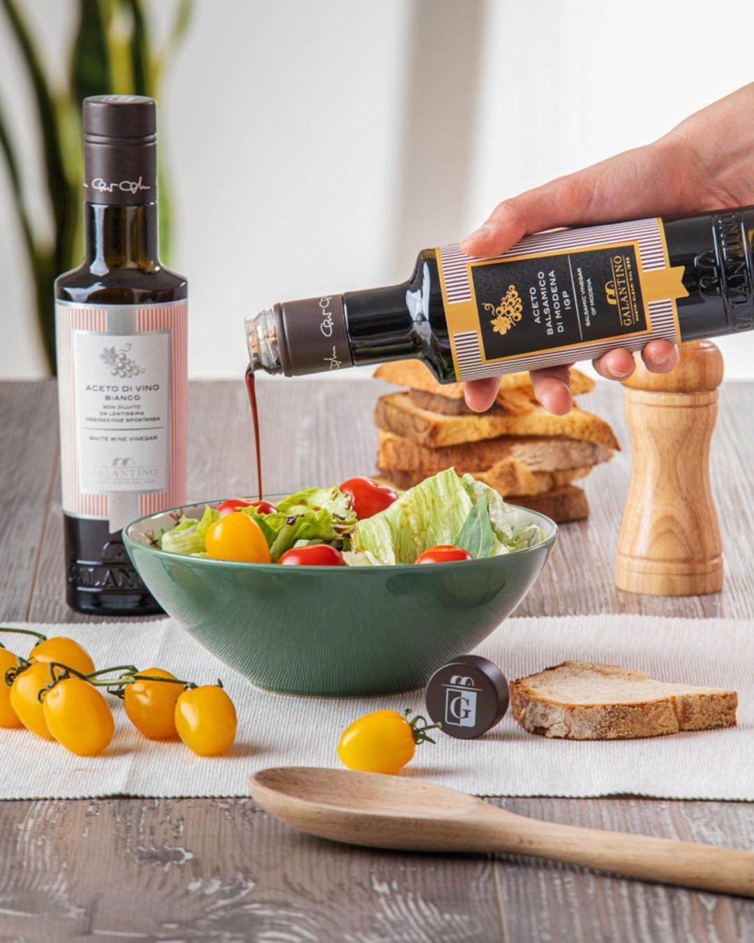 Balsamic Vinegar of Modena PGI being poured over fresh salad in green bowl, with cherry tomatoes and bread on rustic table