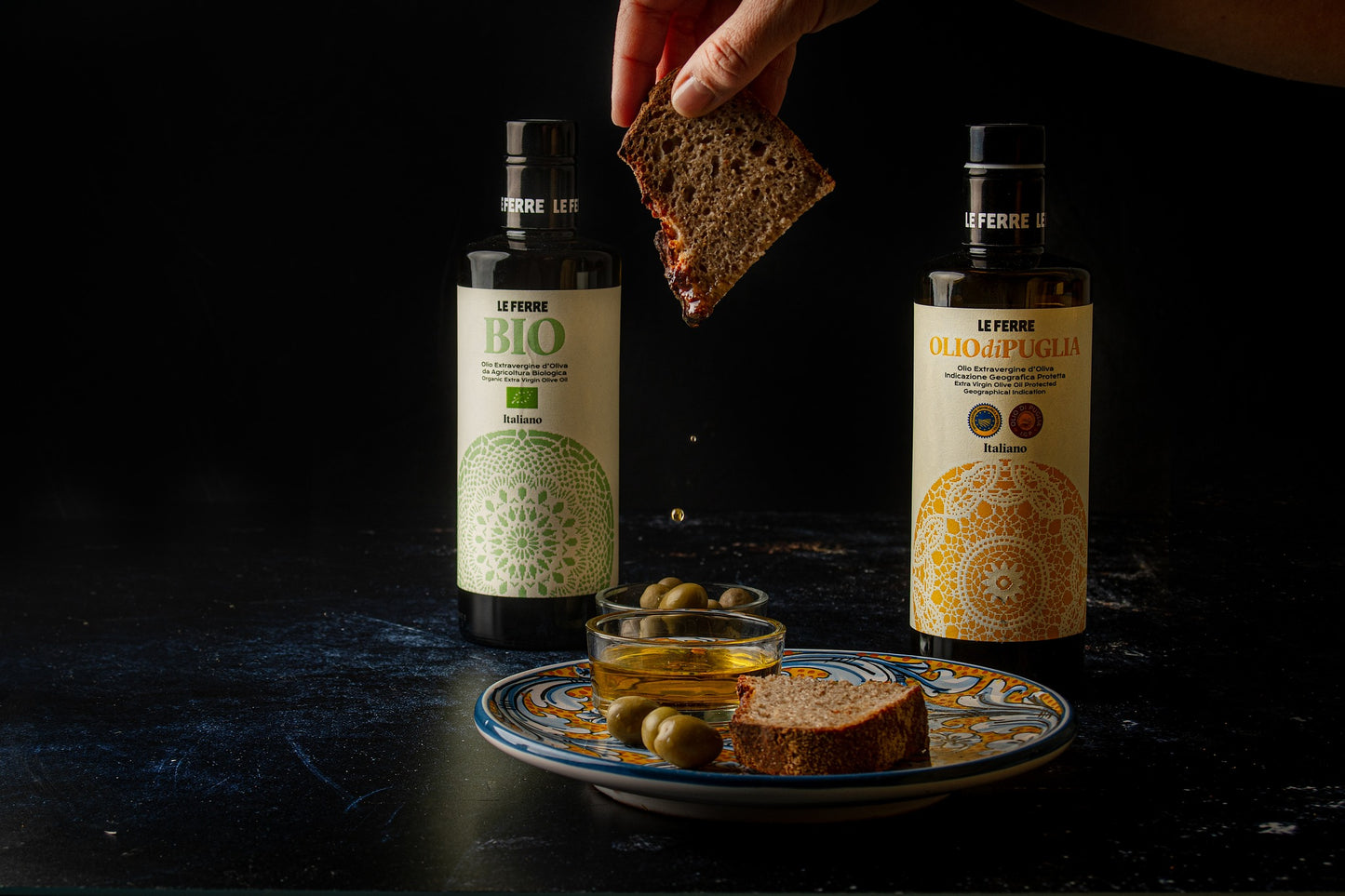 LeFerre Olio di Puglia IGP bottles with bread slice being dipped, served alongside green olives on decorative plate