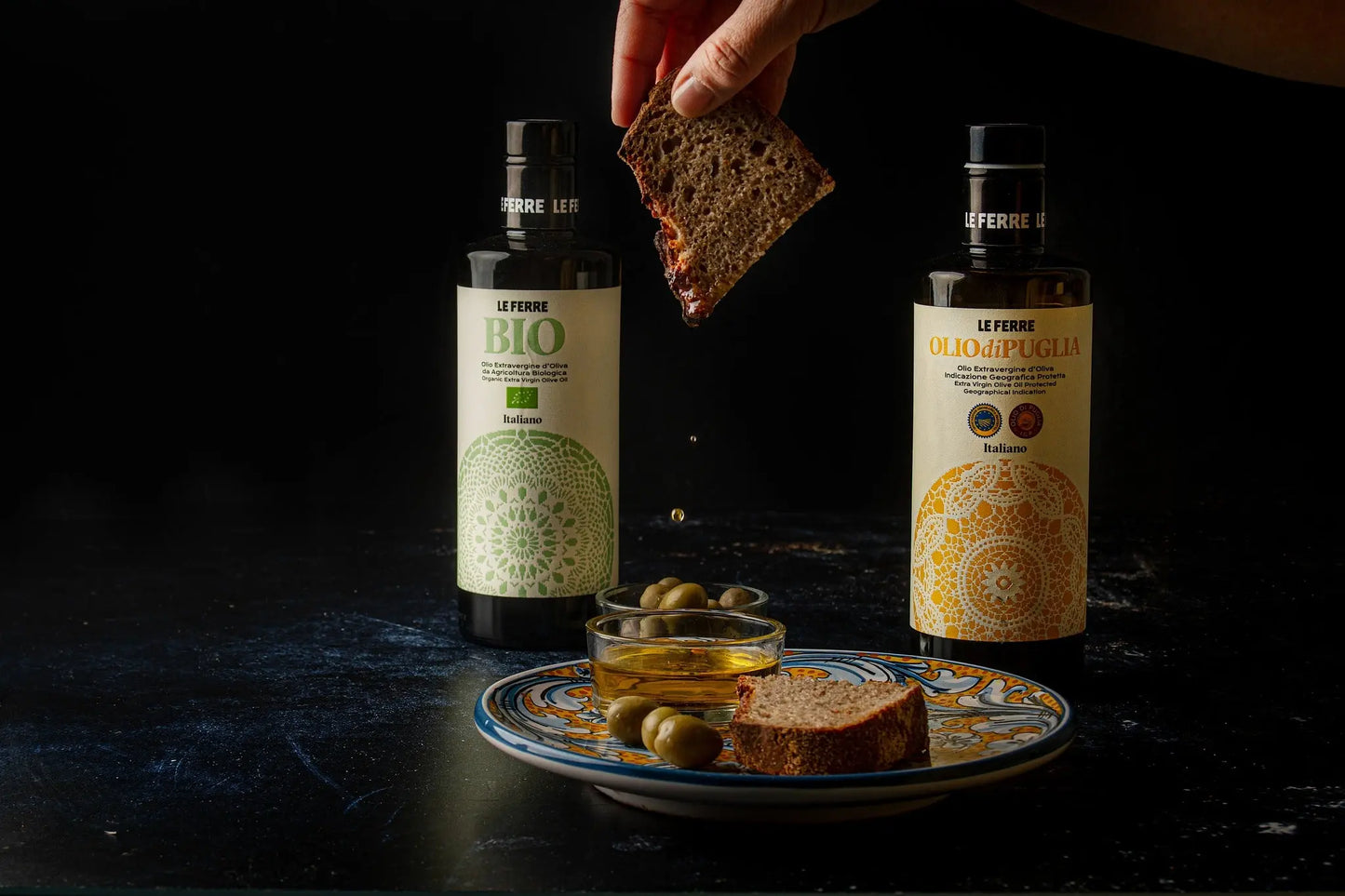 LeFerre Olio di Puglia IGP bottles with bread slice being dipped, served alongside green olives on decorative plate