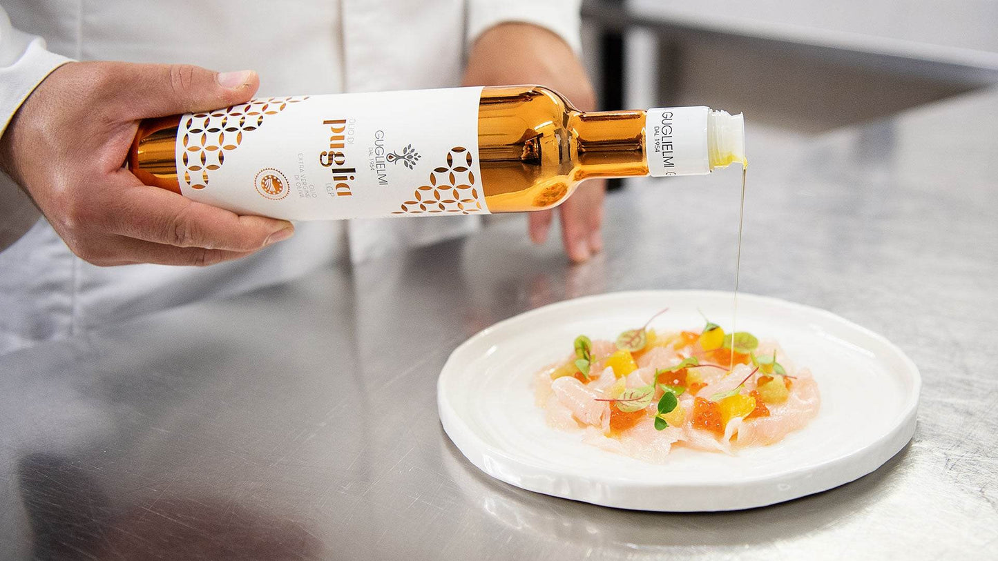 Guglielmi Puglia PGI olive oil being poured over a fresh seafood dish on white plate in professional kitchen setting